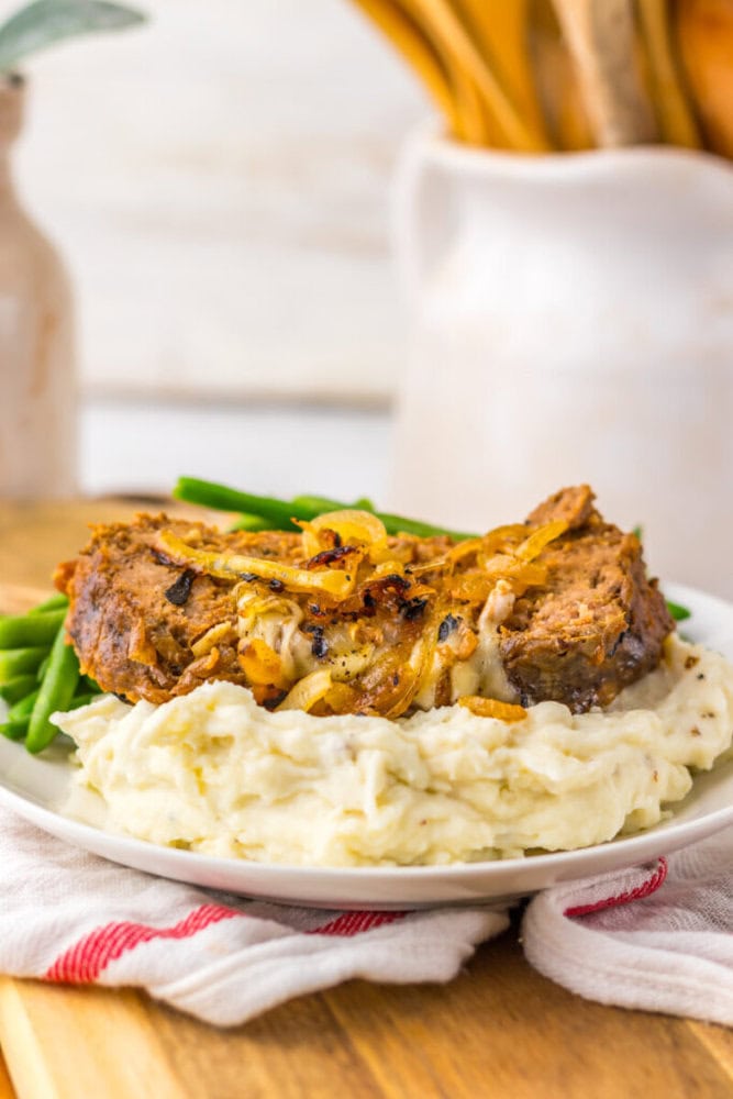 A plate of mashed potatoes topped with a slice of meatloaf and caramelized onions, served with green beans.