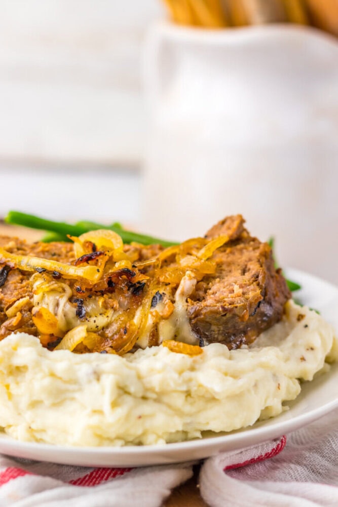 A plate of mashed potatoes topped with a slice of meatloaf and sautéed onions, with green beans in the background.