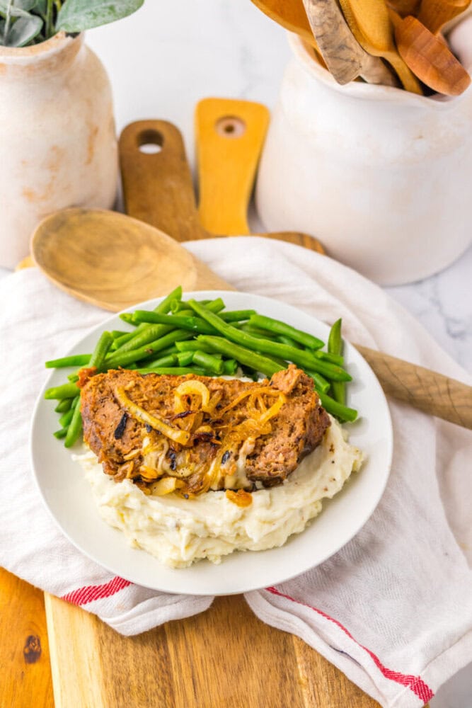 A plate with a slice of meatloaf topped with onions, served over mashed potatoes and green beans, sits on a wooden board with a wooden spoon beside it.