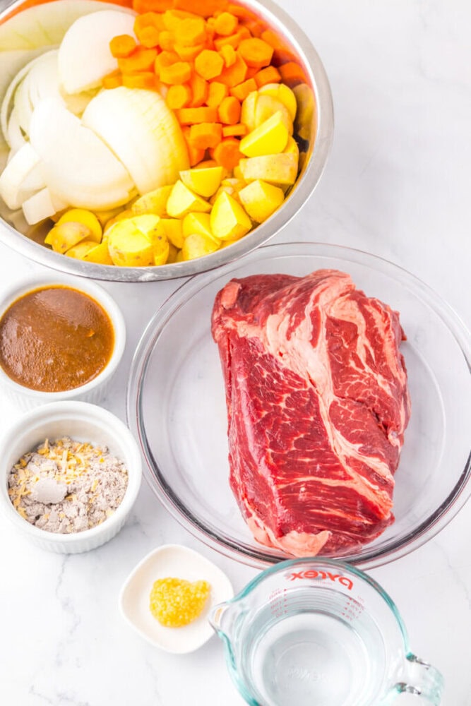 A raw beef roast, sliced onions, carrots, potatoes, broth, seasoning mix, minced garlic, and water are arranged in separate bowls on a white surface.