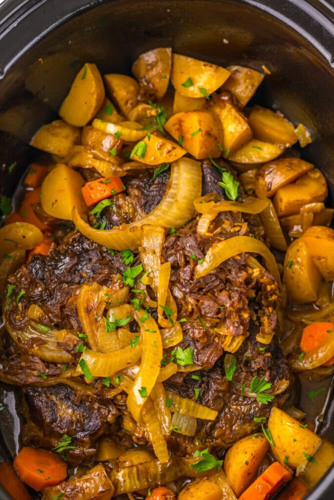 A slow cooker pot roast with sliced onions, carrots, and potatoes, garnished with chopped parsley.