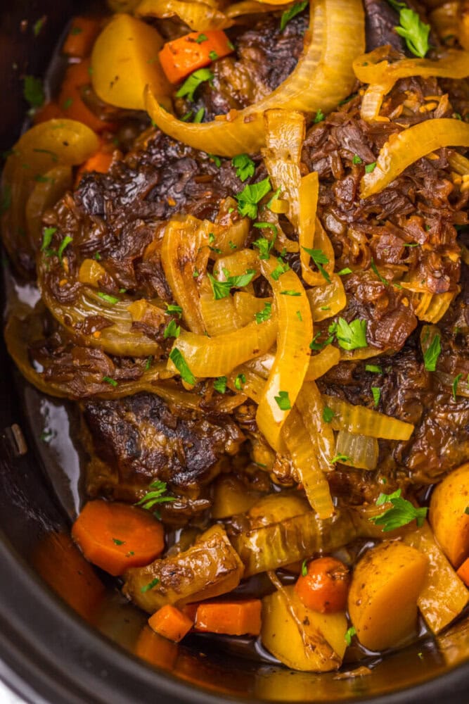 Close-up of a slow-cooked beef pot roast with sliced onions, carrots, and potatoes, garnished with chopped parsley.