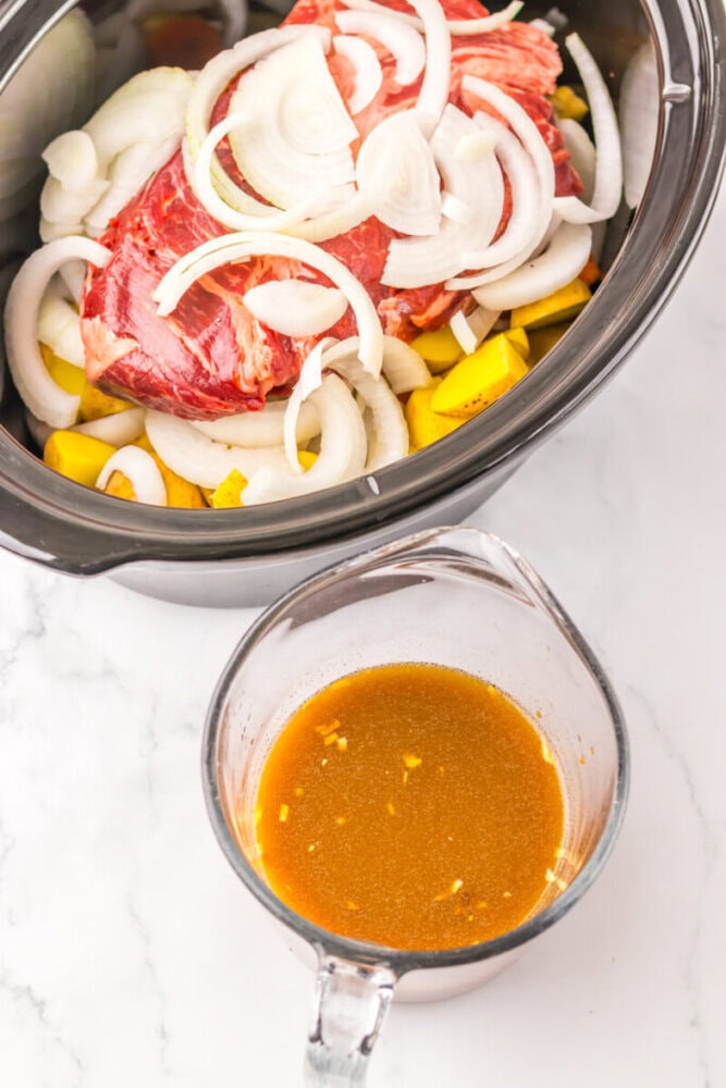 A slow cooker with raw beef, sliced onions, and potatoes, next to a glass measuring cup filled with brown seasoned broth.
