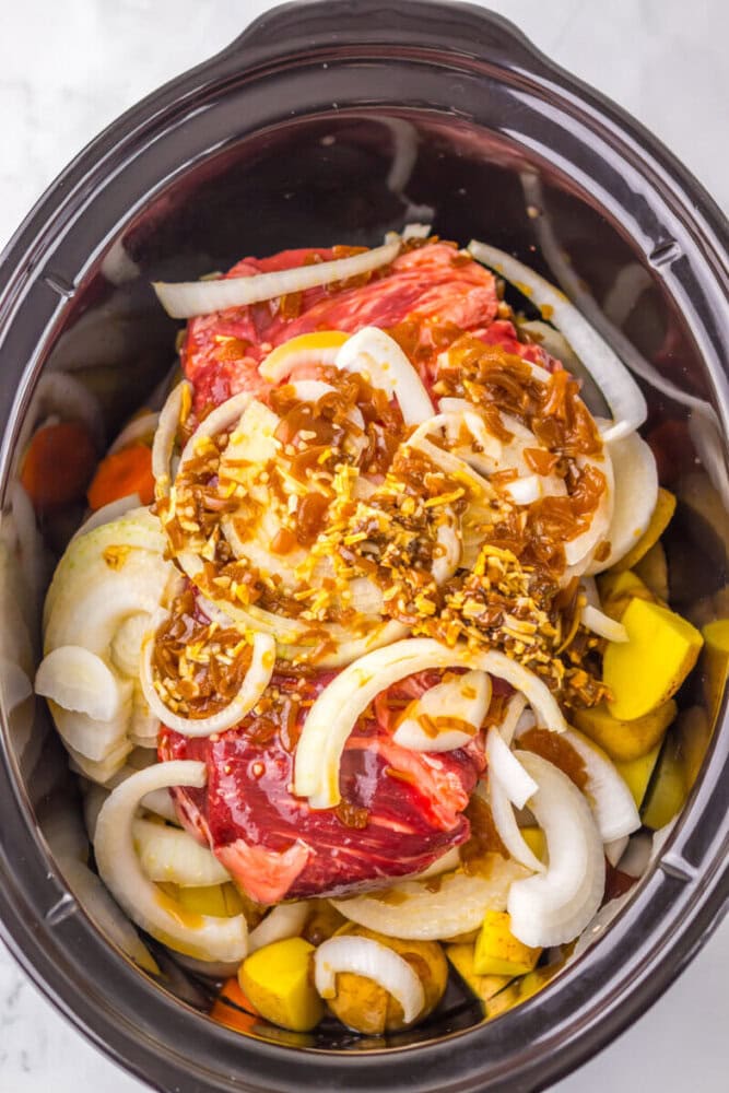 A slow cooker filled with raw beef, sliced onions, chopped vegetables, and a brown seasoning sauce.
