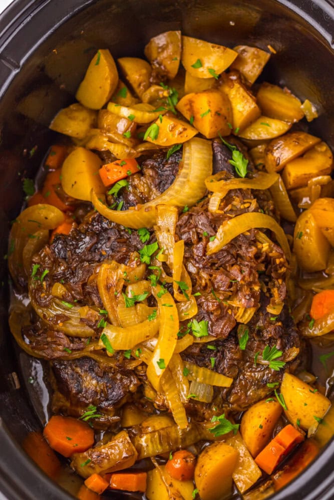 A slow-cooked pot roast topped with sliced onions, surrounded by cooked potatoes and carrots, garnished with chopped parsley.