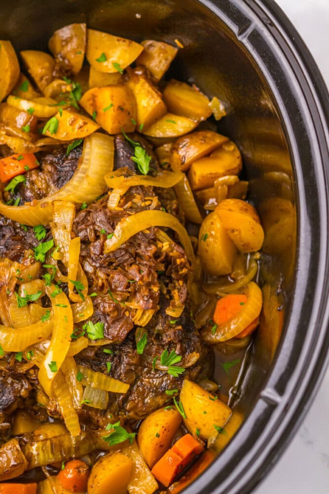 A cooked pot roast with sliced onions, carrots, potatoes, and herbs in a slow cooker.