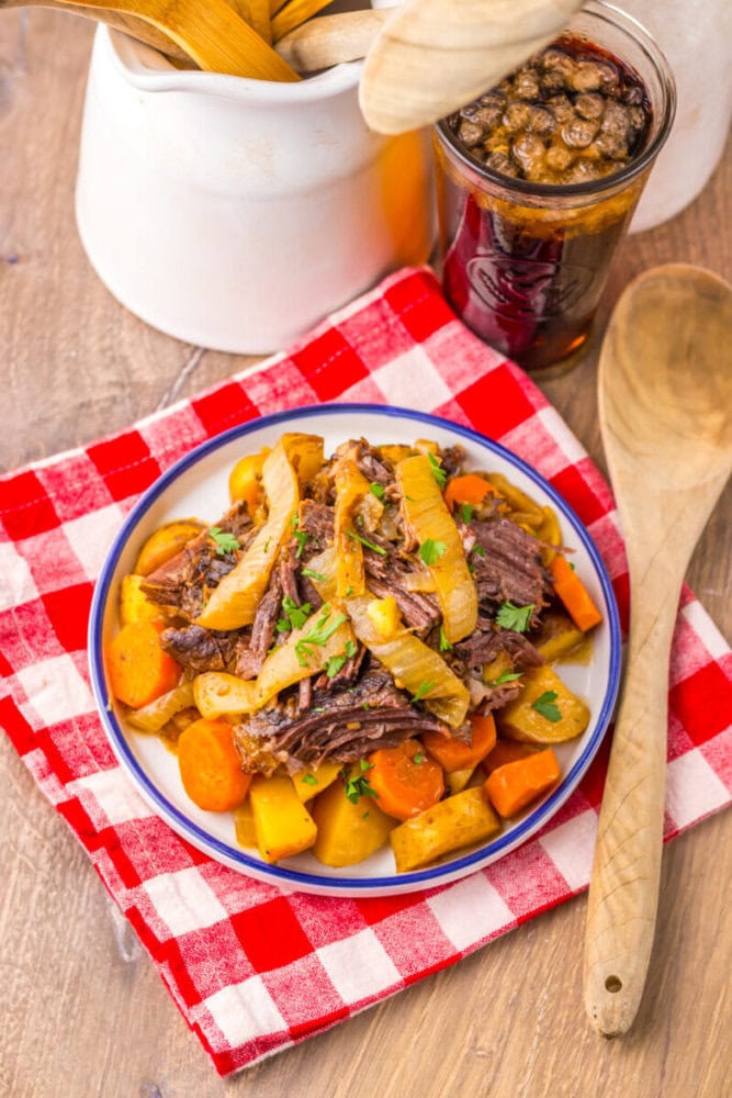 A plate of pot roast with carrots, potatoes, and onions sits on a red checkered napkin beside a wooden spoon and a jar of pickles.