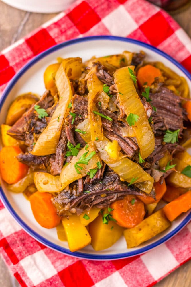 A plate of pot roast with sliced onions, carrots, and potatoes, garnished with parsley, sits on a red and white checkered cloth.