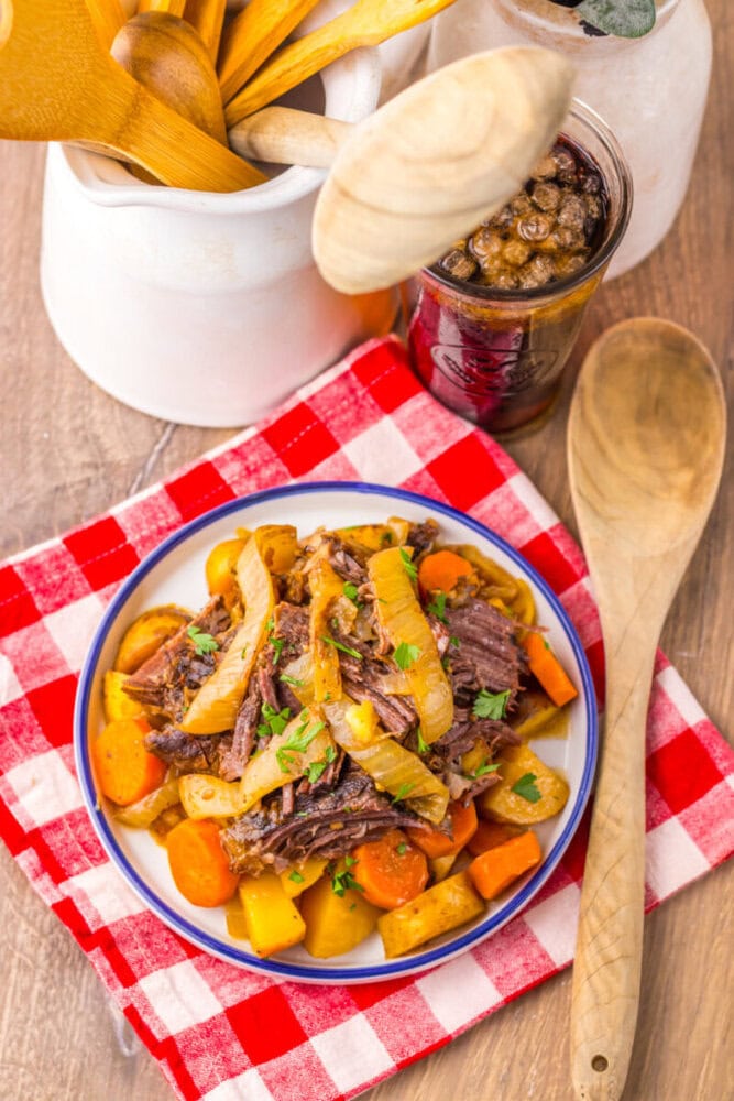 A plate of pot roast with carrots, potatoes, and onions sits on a red checkered napkin, next to a wooden spoon and a container holding wooden utensils.