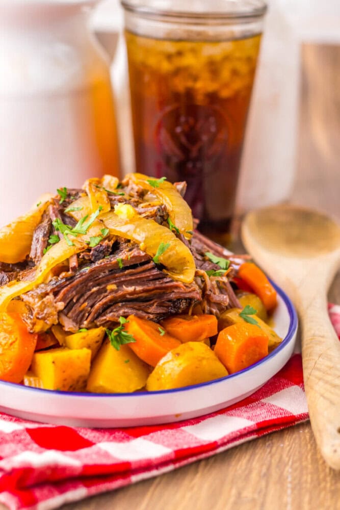A plate of pot roast with sliced beef, carrots, potatoes, and onions, garnished with herbs, sits on a table next to a glass of iced tea and a wooden spoon.