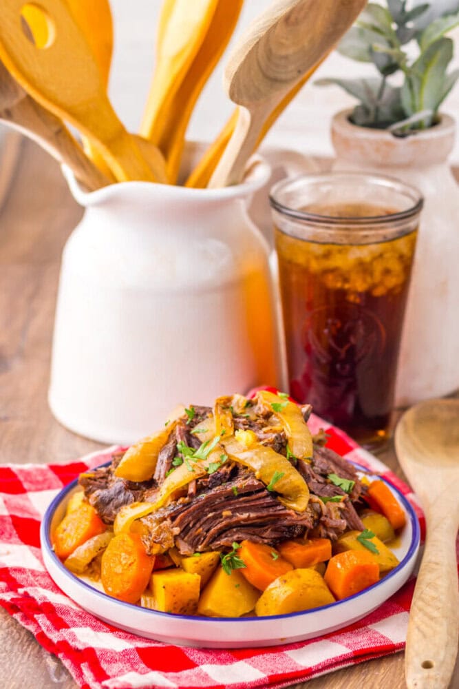 A plate of pot roast with carrots and potatoes sits on a red checkered napkin, next to a glass of iced drink and a white container holding wooden utensils.