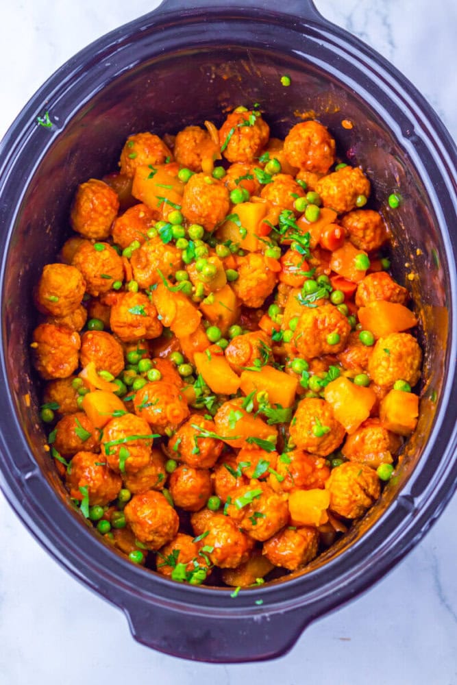 Slow cooker filled with meatballs, diced potatoes, green peas, and sauce, garnished with chopped herbs.