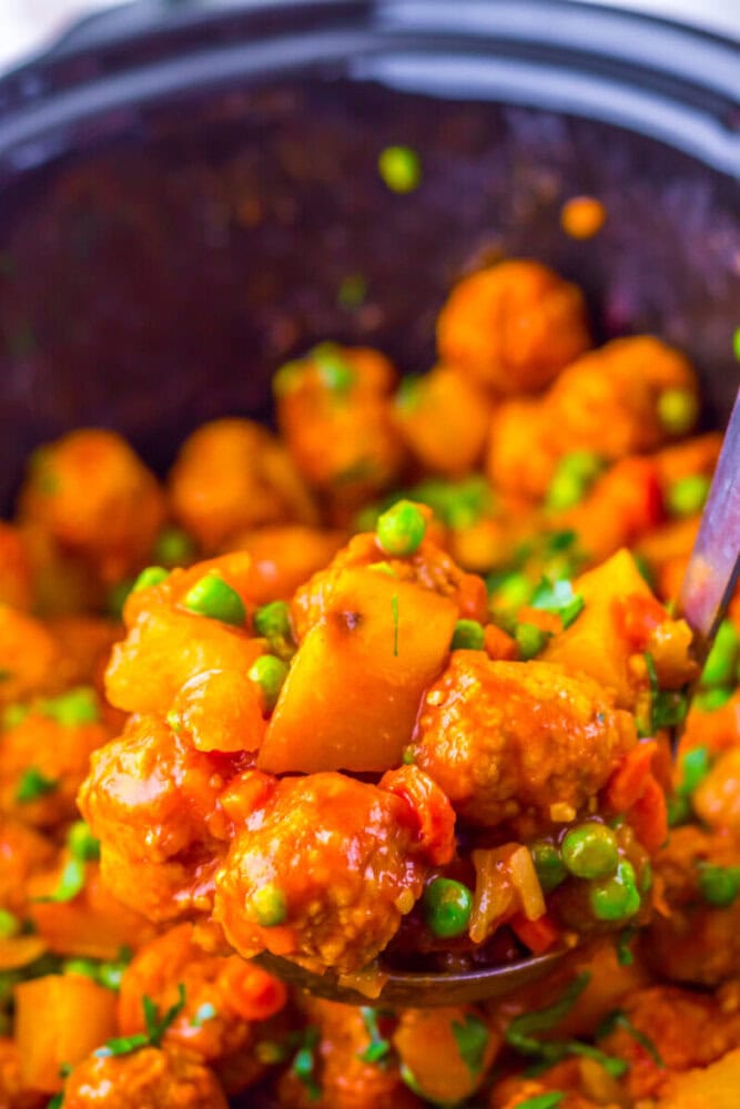 Meatballs cooked in a tomato-based sauce with peas, diced potatoes, and carrots, served from a pot with a ladle.