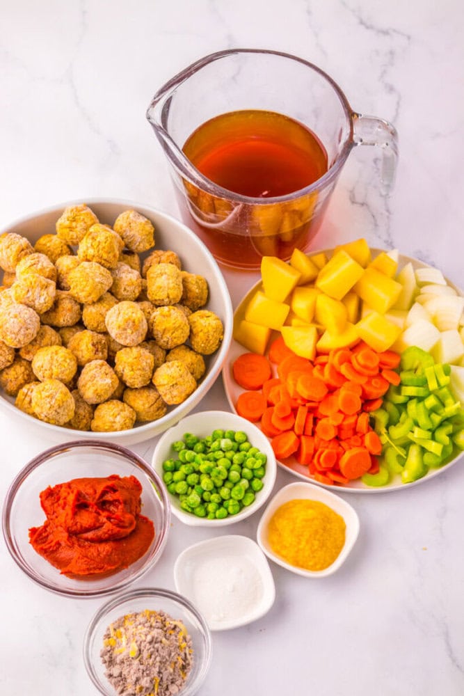 Ingredients for soup displayed on a white surface, including broth, meatballs, diced potatoes, carrots, celery, onions, peas, tomato paste, and seasonings in separate bowls.