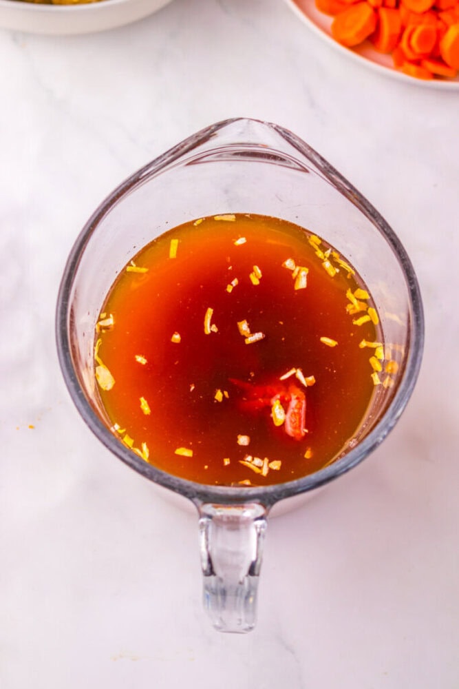 A glass measuring cup filled with brown broth containing chopped onions and tomatoes, placed on a white surface.