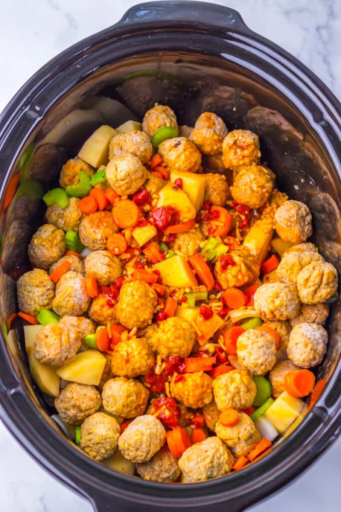 A slow cooker filled with meatballs, diced potatoes, carrots, celery, and sauce, ready to be cooked.