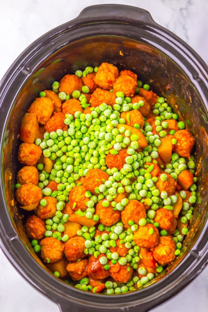 Slow cooker filled with meatballs, diced potatoes, sliced carrots, and topped with a layer of frozen peas.