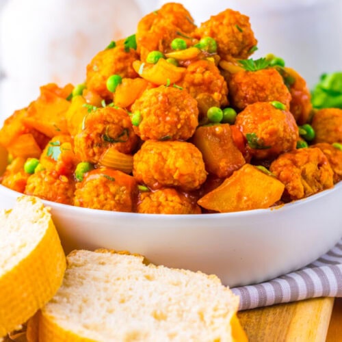 A white bowl filled with saucy meatballs, peas, and potatoes, served with two slices of bread on a wooden board.