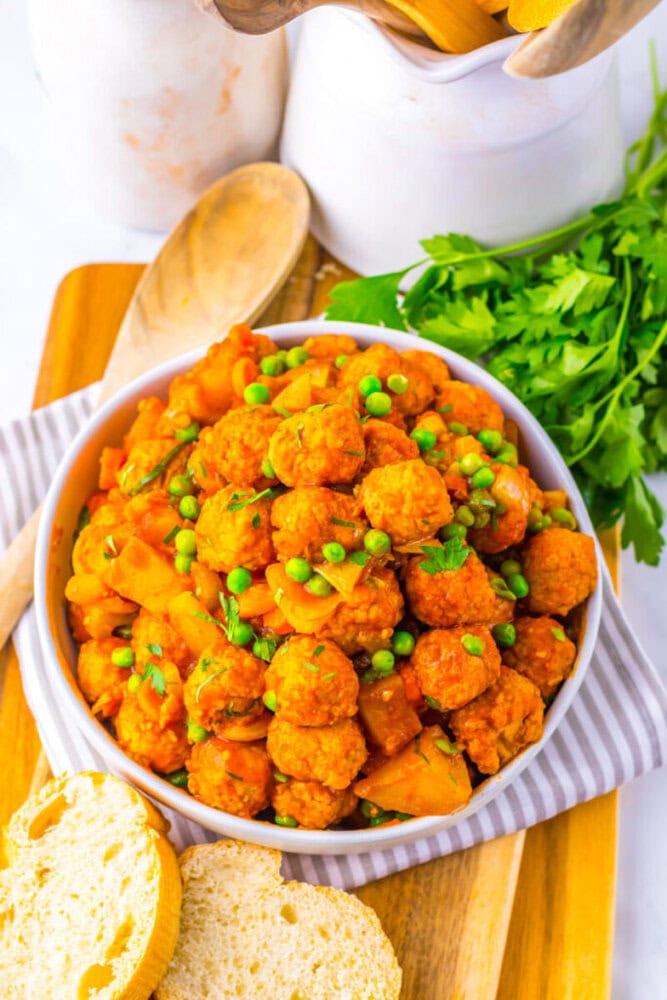 A bowl of vegetable stew with peas and meatballs, garnished with parsley, placed on a wooden board with slices of bread beside it.