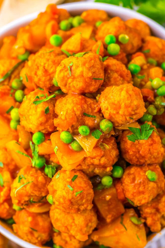 A close-up of a bowl filled with vegetable curry featuring round kofta balls, green peas, diced potatoes, and garnished with fresh herbs.