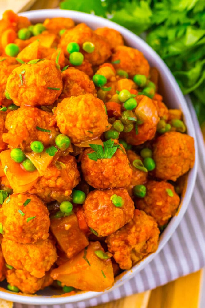 A close-up of a bowl filled with vegetable and pea curry featuring round, orange-colored vegetable balls and chopped greens, placed on a striped cloth.