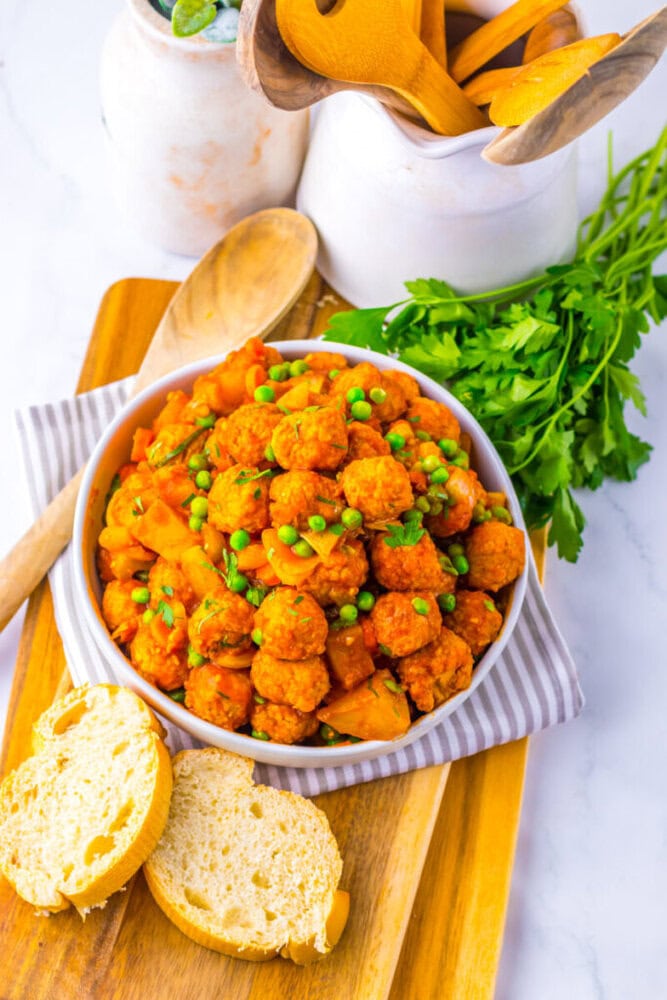 A bowl of meatballs with peas and diced potatoes in tomato sauce, garnished with parsley, sits on a striped cloth next to sliced bread and wooden utensils.