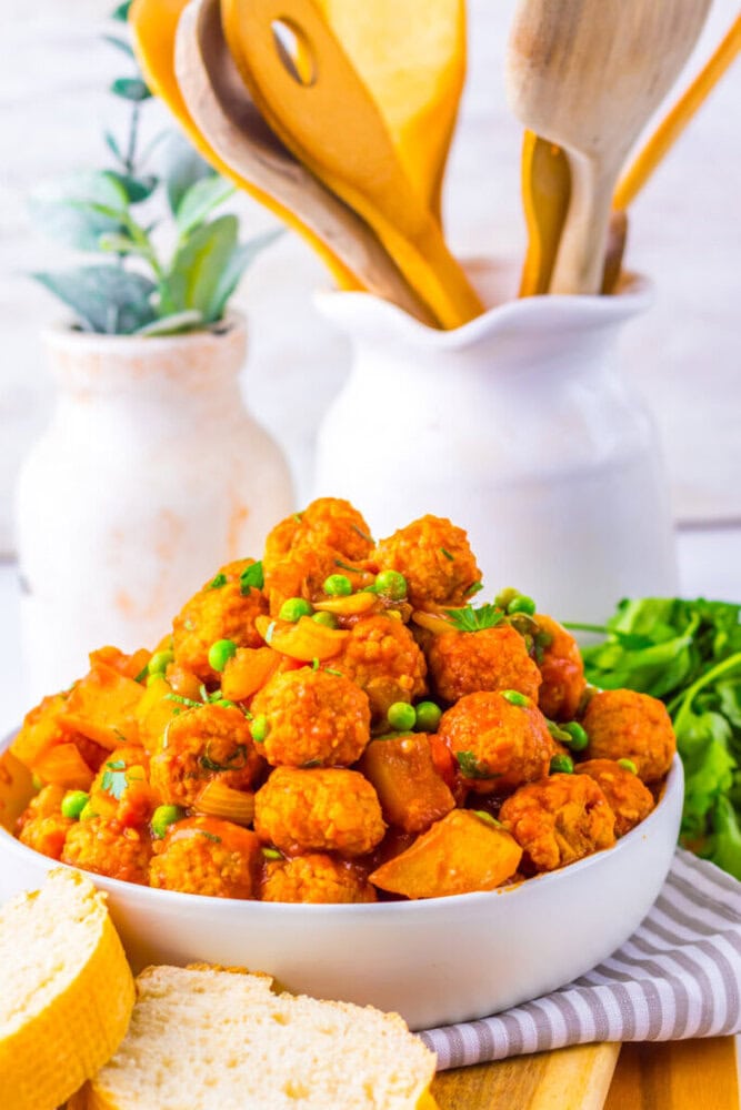 A white bowl filled with meatballs in red sauce with peas and potatoes, placed on a table alongside bread slices and wooden utensils in the background.