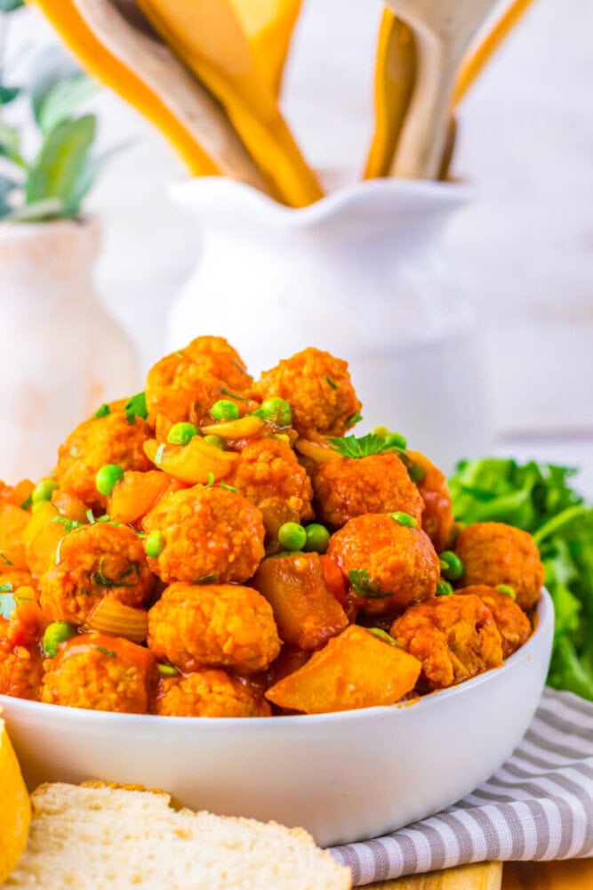 A white bowl filled with vegetable meatballs in red sauce, mixed with peas and potatoes, placed on a striped cloth with kitchen utensils in the background.