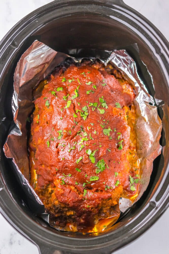 Meatloaf with tomato glaze topped with chopped parsley sits in a slow cooker lined with aluminum foil.