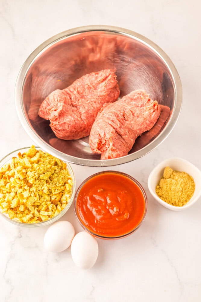 A metal bowl with raw ground meat, next to bowls of bread crumbs, tomato sauce, grated cheese, and two eggs on a white surface.