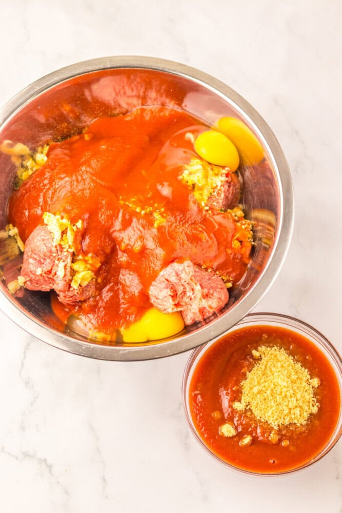 A metal bowl with ground meat, eggs, tomato sauce, and seasoning; next to a smaller bowl with tomato sauce and seasonings on a white surface.