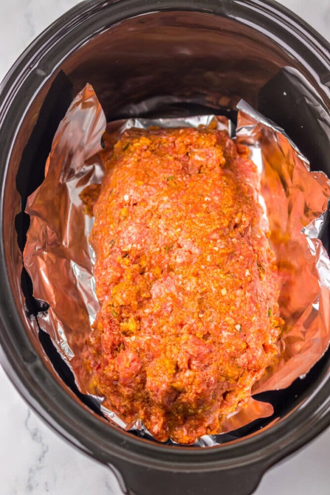 Uncooked meatloaf placed on aluminum foil inside a slow cooker, ready for cooking.