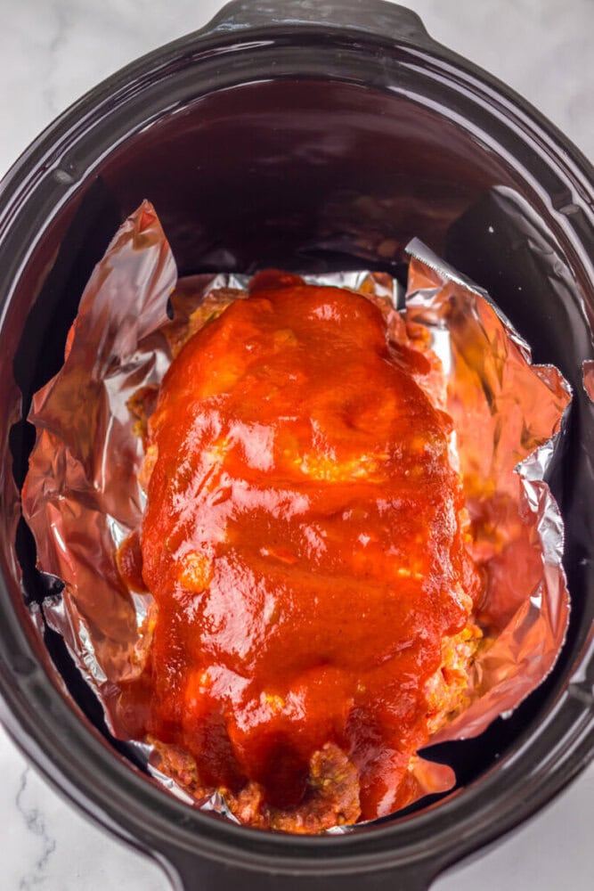 A meatloaf covered in red sauce sits on aluminum foil inside a slow cooker.