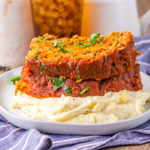 Two slices of meatloaf topped with tomato sauce and parsley rest on a serving of mashed potatoes, on a white plate with a blue striped cloth underneath.