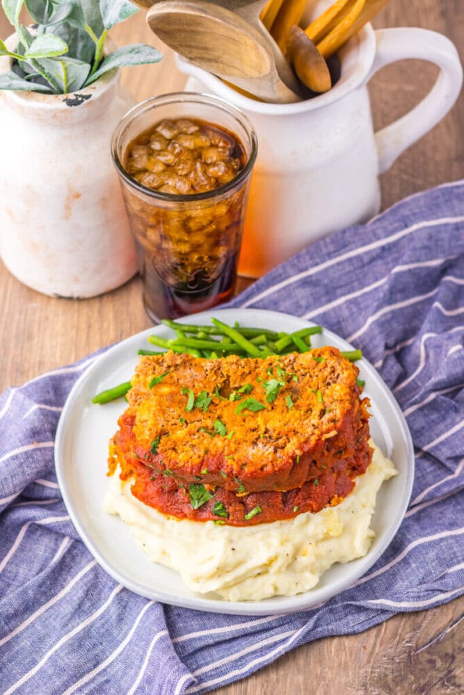 A plate of meatloaf topped with tomato sauce and herbs sits on mashed potatoes, with green beans on the side and a glass of iced cola in the background.