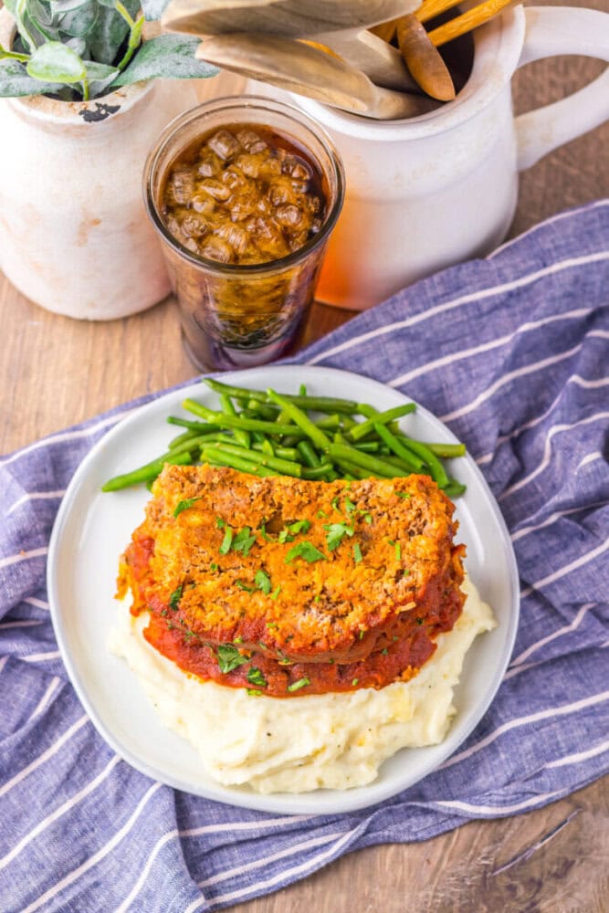 A plate with two slices of meatloaf on mashed potatoes, green beans on the side, and a glass of iced drink on a wooden table with a blue striped cloth.