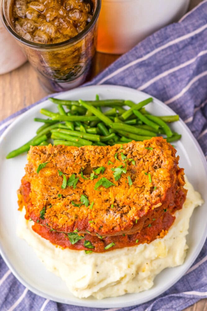 Two slices of meatloaf topped with parsley sit on mashed potatoes, with green beans on the side, next to a glass of soda on a striped cloth.