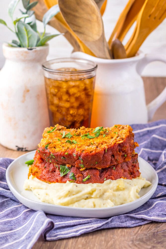 A plate with two slices of meatloaf topped with herbs, served on mashed potatoes, sits in front of a glass of iced drink and kitchen utensils.