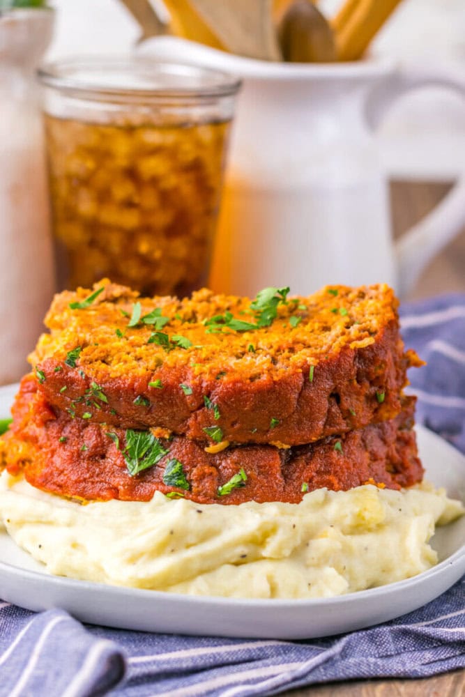 Two slices of meatloaf topped with tomato sauce and parsley are served on a bed of mashed potatoes, with a glass of iced tea and kitchen utensils in the background.