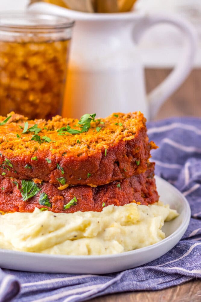 A slice of meatloaf with tomato sauce sits on top of a serving of mashed potatoes, garnished with chopped parsley, with a drink in the background.