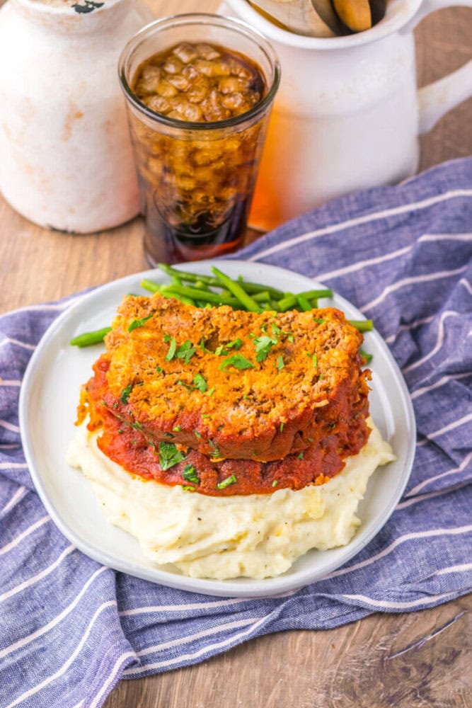 A plate with two slices of meatloaf on mashed potatoes, topped with tomato sauce and parsley, green beans on the side, and a glass of soda in the background.