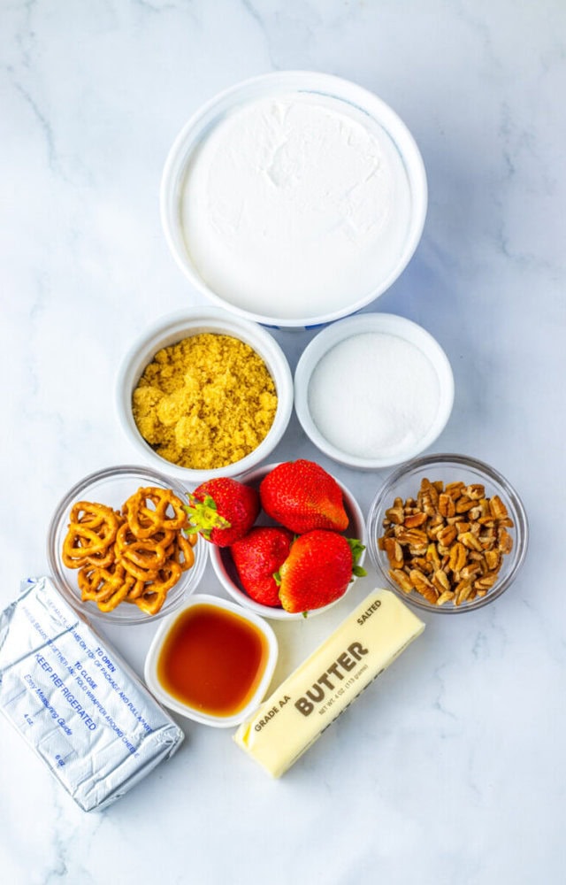 Seven small containers holding cream cheese, whipped topping, brown sugar, granulated sugar, pretzels, strawberries, chopped pecans, butter, and vanilla extract on a white surface.