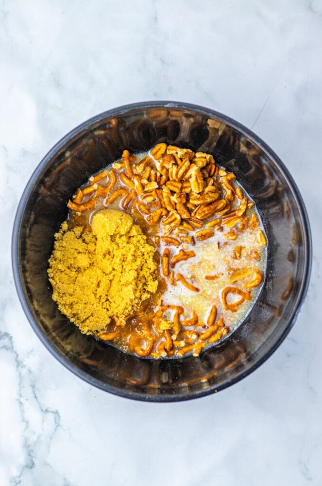 A black bowl containing crushed pretzels, brown sugar, and a liquid mixture on a white marble surface.