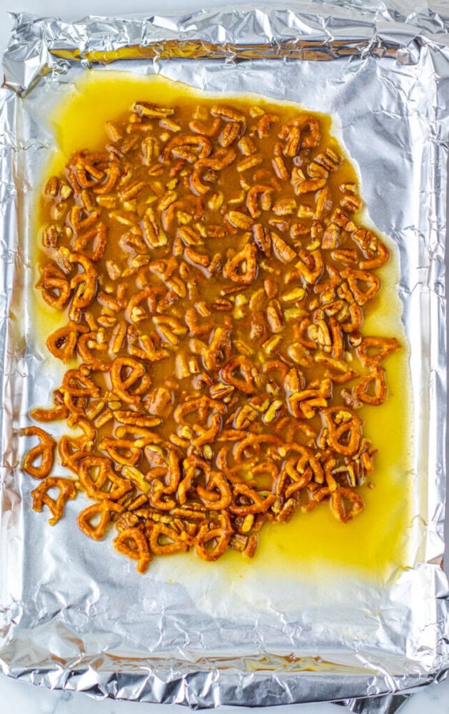 Broken pretzels coated in a yellow buttery mixture are spread on a foil-lined baking sheet.