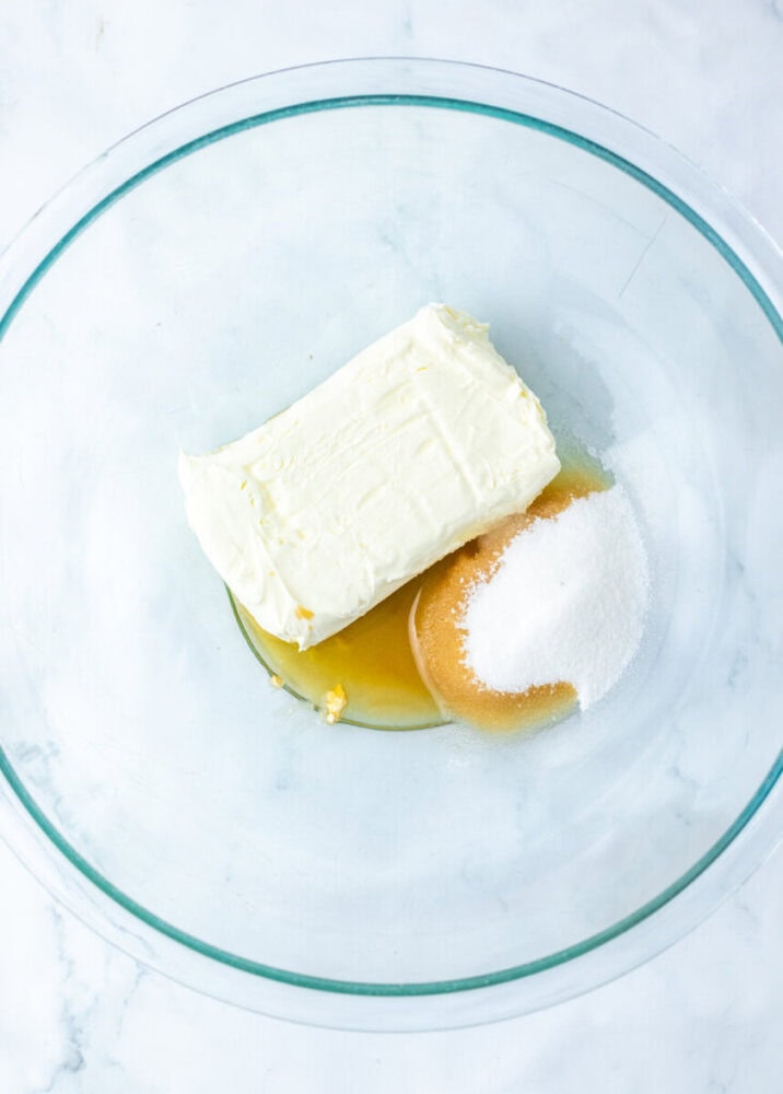 A glass mixing bowl containing a stick of butter, brown sugar, granulated sugar, and a splash of vanilla extract on a white surface.