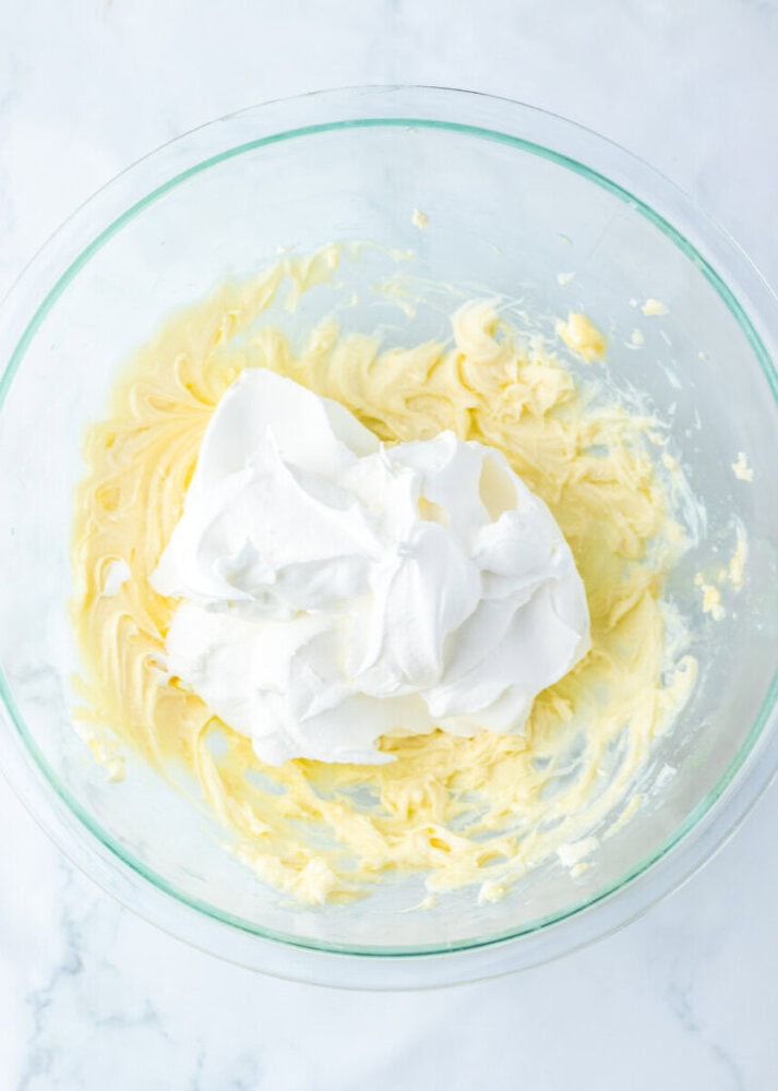 A glass bowl containing partially mixed yellow batter and a dollop of whipped cream or meringue on top, on a white marble surface.