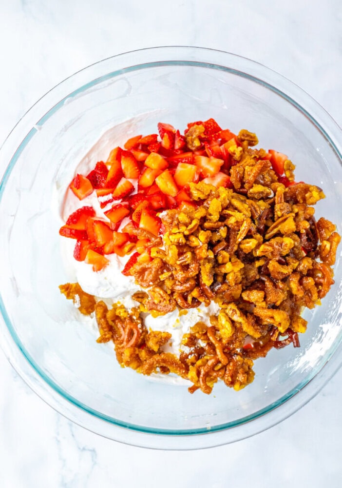 A glass bowl containing chopped strawberries, cream cheese mixture, and candied pretzel pieces on a white surface.