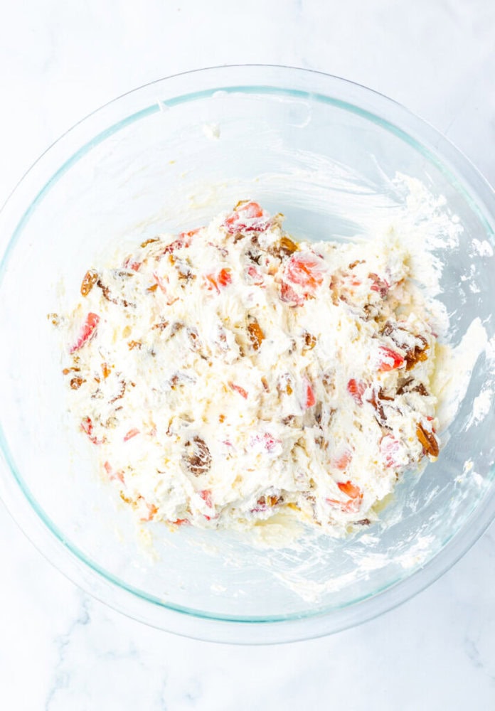 A glass bowl containing a mixture of whipped cream, chopped strawberries, and chopped nuts on a white marble surface.