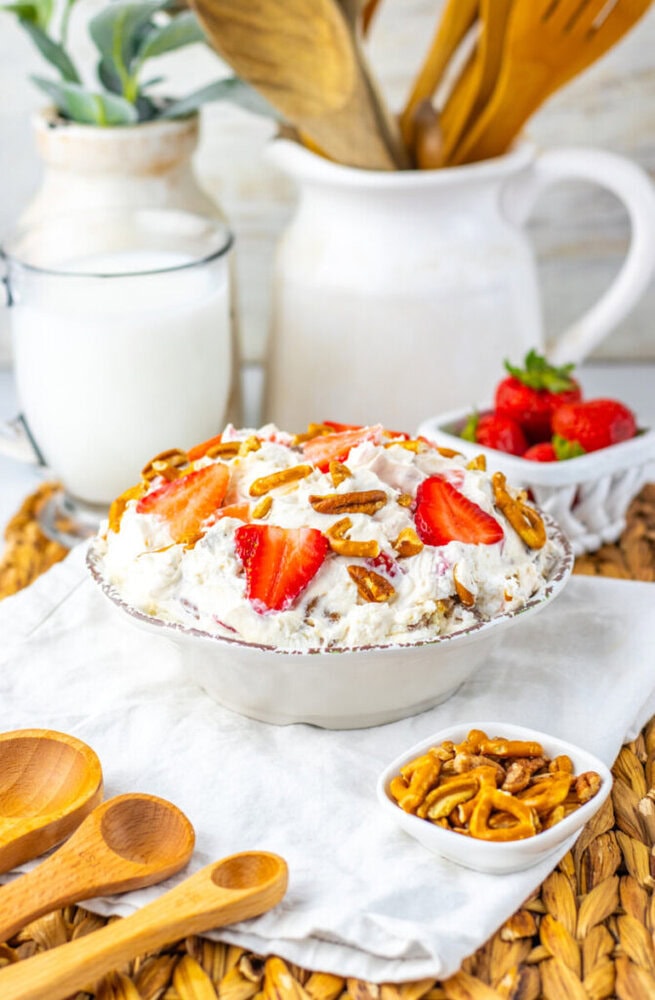 A bowl of creamy dessert topped with sliced strawberries and pretzel pieces, with a glass of milk, fresh strawberries, and wooden utensils in the background.