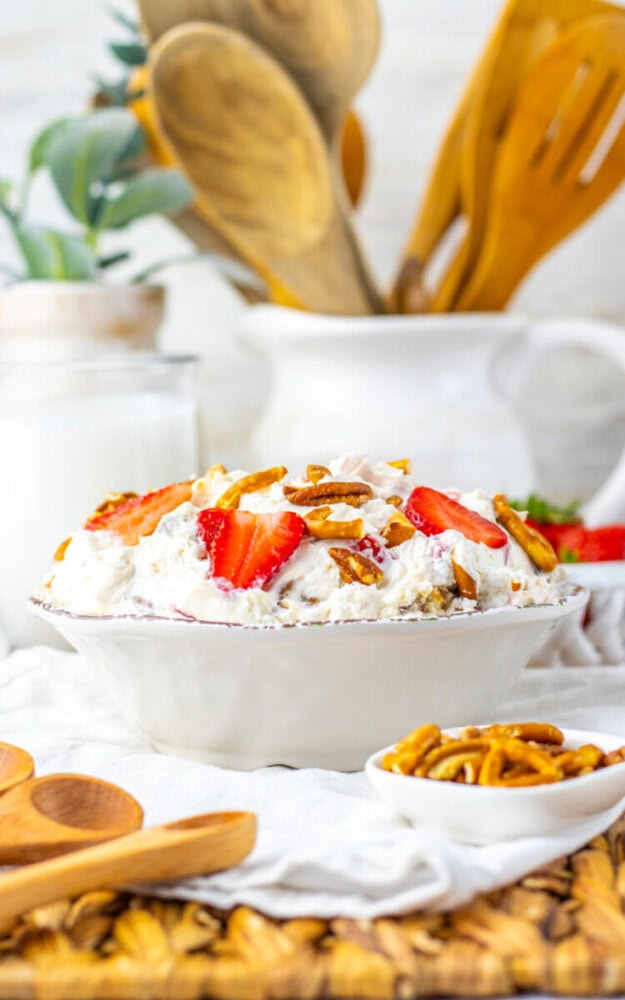 A bowl of creamy dessert topped with sliced strawberries and pretzel pieces, placed on a white cloth with wooden utensils and a jar in the background.